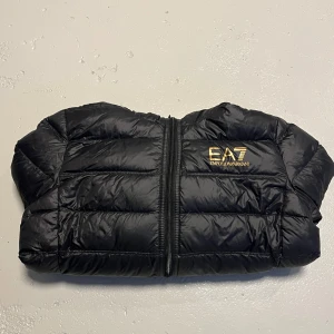 Svart pufferjacka från Emporio Armani - Säljer en snygg svart pufferjacka från Emporio Armani med EA7-logga i guld på bröstet. Jackan har en dragkedja framtill och en praktisk huva. Perfekt för kyliga dagar!