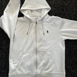Vit hoodie från Polo Ralph Lauren - Säljer en stilren vit hoodie från Polo Ralph Lauren med dragkedja och klassisk logga på bröstet. Tröjan har en justerbar huva med snören och praktiska fickor framtill. Perfekt för en avslappnad look.
