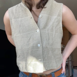 Beige stickad väst från Kathleen Madden NY - Säljer en stilren beige stickad väst från Kathleen Madden NY. Västen har en v-ringad design med knappar framtill, perfekt för lager-på-lager-stil. Ett mångsidigt plagg för olika tillfällen.