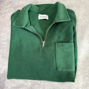 Grön tröja med dragkedja från Woodbird - Säljer en snygg grön tröja från Woodbird med dragkedja och krage. Tröjan har en praktisk bröstficka och långa ärmar. Perfekt för en stilren och avslappnad look.