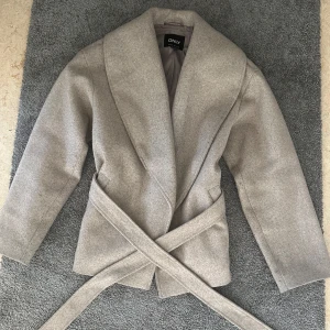 Beige kappa från ONLY - Snygg beige kappa från ONLY med ett elegant bälte i midjan. Jackan har en stilren design med långa ärmar och en mjuk krage. Perfekt för kyligare dagar, men även nu under våren🌺