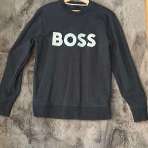Svart sweatshirt från Boss - Snygg svart sweatshirt från Boss med stort vitt tryck på framsidan. Tröjan har långa ärmar och ribbade muddar vid ärmslut och nederkant. Perfekt för en stilren och avslappnad look. Hör av er för frågor 🥰