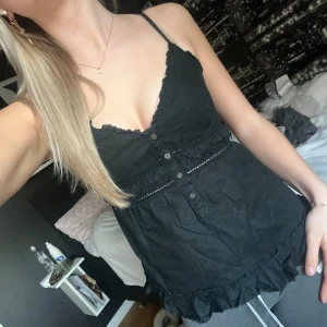 Svart ärmlös topp med spetsdetaljer - Snygg svart ärmlös topp med smala axelband och spetsdetaljer. Toppen har knappar framtill och en volangkant nedtill. Knytning där bak. 