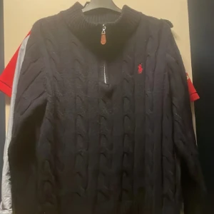 marinblå stickad tröja från Ralph Lauren - Säljer en marinblå stickad tröja från Ralph Lauren med dragkedja vid halsen och ett rött broderat märke på bröstet. Perfekt för kyliga dagar med sin kabelstickade design och höga krage.