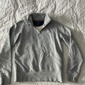 Grå halvzip tröja från GANT - Säljer en stilren grå halvzip tröja från GANT i storlek XS. Tröjan har hög krage med dragkedja och diskret logga på bröstet. Perfekt för lager-på-lager och passar till många olika outfits.