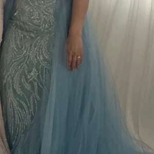 Ljusblå långklänning med glitterdetaljer - Säljer en ljusblå långklänning med glittriga mönster och voluminöst tyllsläp. Klänningen har figurnära passform och är perfekt för bal eller fest. Klänning är i nyskick i st 36/38 med avtagbart släp. 