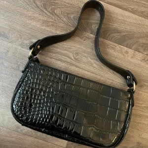 Svart axelväska - Säljer en svart axelväska med glansig yta. Väskan har en bred axelrem och guldfärgade metalldetaljer. Perfekt för att lyfta vilken outfit som helst. säljer den för 160kr