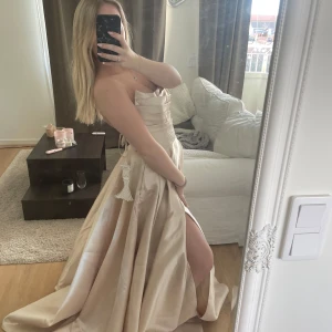 Champagnefärgad långklänning med slits - OANVÄND!! Lappar kvar. Säljer en elegant axelbandslös långklänning i champagnefärgat satinliknande material. Klänningen har en hög slits framtill och markerad midja med dekorativ knytning. Perfekt för bal eller fest. Snygg och tidlös design.