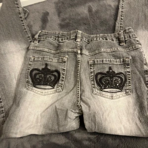 Grå jeansbyxor med broderad krona - Säljer ett par grå jeansbyxor från Tutti Frutti med coola broderade kronor på bakfickorna. Byxorna har en klassisk femficksmodell och raka ben. Perfekta för dig som vill sticka ut lite extra med detaljer på jeansen. Dom har tappat några stenar på bak fickorna🤍
