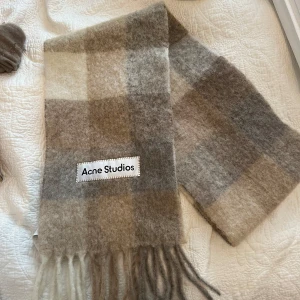 Rutig halsduk från Acne Studios - Säljer en supermjuk och stilren rutig halsduk från Acne Studios. Den har en mix av beige och grå toner med fransar i ändarna. Perfekt för att hålla dig varm och snygg under kyliga dagar.