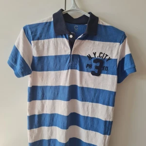 Blåvit randig pikétröja från Cubus (barn) - Säljer en snygg blåvit randig pikétröja med korta ärmar och krage. Tröjan har en broderad text 'N.Y. City' och siffran '3' på bröstet. 100% bomull. Storlek 158/164. Kan hämtas i Vallentuna.