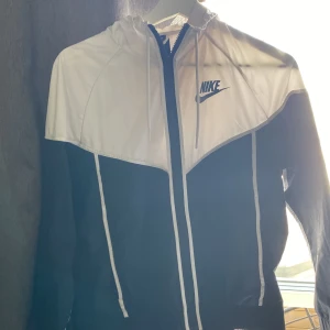 Svart och vit vindjacka från Nike - Nike vindjacka i svart och vitt med huva och dragkedja framtill. Jackan har lång ärm och sportig design med kontrasterande detaljer och Nike-logga på bröstet. Perfekt för utomhusaktiviteter och enkel att matcha med olika outfits. Kolla in min profil för mer
