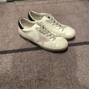 Vita sneakers med stjärndetalj från Golden Goose - Säljer ett par vita sneakers från Golden Goose med grå snörning, silverfärgad stjärna på sidan och svart hälparti. Klassisk rund tå och diskret logga på sidan. Perfekta för en stilren och trendig look.