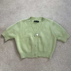 Ljusgrön ribbad kofta från SHEIN - Kort, ljusgrön kofta från SHEIN med ribbad struktur och v-ringning. Koftan har korta ärmar och knäpps framtill med vita knappar. Perfekt att slänga över en klänning eller topp för en chill look.