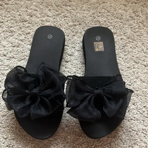 Svarta sandaler med stor rosett - Säljer ett par svarta sandaler med en stor, dekorativ rosett framtill. Skorna har platt sula och öppen tå, perfekta för varma dagar. rosetten är i ett tunt, genomskinligt tyg.