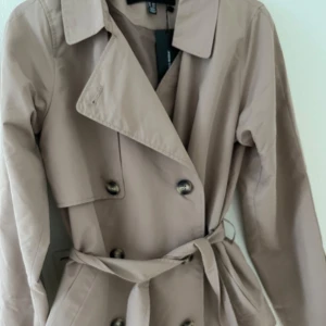 Beige trenchcoat från H&M - Snygg beige trenchcoat från H&M med klassisk design. Jackan har knappar framtill och ett bälte i midjan för en stilren look. Perfekt för vår och höst! Säljer en helt ny trenchcoat, storlek XS (något stor i storleken). Aldrig använd, endast testad - lappar kvar! Superfin men kommer tyvärr inte till användning.  Köpare står för frakt.