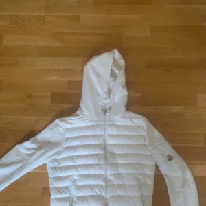 Vit quiltad jacka från Moncler - Säljer en snygg vit quiltad jacka från Moncler med dragkedja och huva. Jackan har ett stilrent märke på ärmen och är perfekt för kyligare dagar. Den är både praktisk och trendig, idealisk för dig som vill hålla stilen under våren och sommaren. Möts bara upp i Stockholm eller Uppsala. 