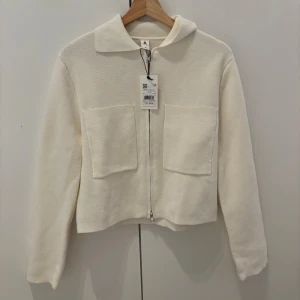 Krämvit stickad kofta med dragkedja - Säljer en krämvit stickad kofta från Åhlens med två stora fickor framtill och dragkedja. Koftan har en klassisk krage och långa ärmar. Perfekt för lager-på-lager och enkel att matcha med olika outfits. Helt ny oanvänd med prislapp.