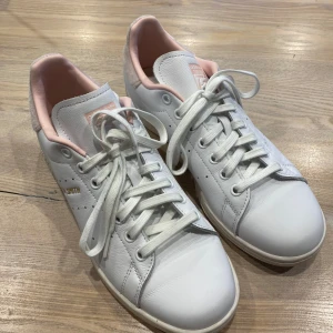 Adidas Stan Smith vita sneakers med rosa detaljer - Säljer ett par fräscha Adidas Stan Smith sneakers i vitt skinn med ljusrosa detaljer och guldbroderad logga på hälen. Klassisk modell med snörning och rund tå. Perfekta för dig som gillar stilrena och tidlösa sneakers. De har varit använda en gång ute men tvättas därefter.🥰
