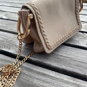 Beige axelväska med guldkedja från H&M - Säljer en beige axelväska från H&M i mjuk mockaliknande material. Väskan har guldfärgade detaljer, kedjerem och dekorativ flätad kant. Perfekt storlek. Den har även två fack inuti.