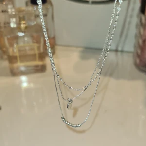 Silverfärgat lager-på-lager halsband - Säljer ett trendigt silverfärgat halsband med flera kedjor i olika längder. Halsbandet har en liten rund berlock och dekorativa pärlor på en av kedjorna. Perfekt för att skapa en snygg lager-på-lager-look💕