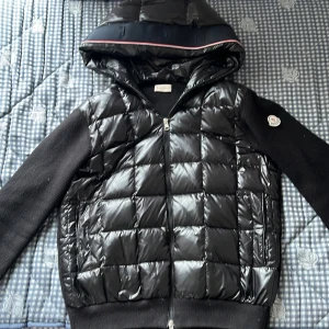 Svart pufferjacka från Moncler med huva - Säljer en svart pufferjacka från Moncler med glansig, quiltad framsida och stickade svarta ärmar. Jackan har huva, dragkedja framtill och Moncler-logga på ärmen. Perfekt för kyliga dagar och riktigt snygg streetstil.
