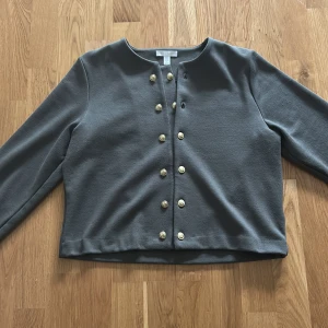Grön cardigan jacka med guldfärgade knappar från H&M - Säljer en stilren grå cardigan jacka från H&M med dekorativa guldfärgade knappar framtill. Jackan har rund halsringning och långa ärmar, perfekt för lager-på-lager-stil.