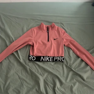Rosa croppad tröja från Nike - Säljer en rosa croppad tröja från Nike med Dri-FIT-teknologi. Tröjan har en halv dragkedja framtill och långa ärmar. Den elastiska nederkanten har texten 'NIKE PRO'. Perfekt för träning eller en sportig look. Aldrig använd fast utan prislapp.