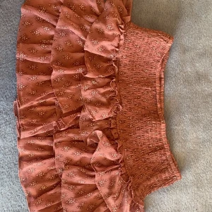 Rosa volangkjol - Charmig rosa kortkjol med volanger och ett diskret blommönster. Kjolen har en elastisk midja och även inbyggda shorts för bekväm passform och är perfekt för varma dagar.