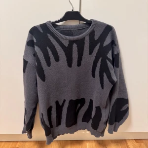 Grå stickad tröja med svart mönster - Säljer en grå stickad tröja med svart abstrakt mönster. Tröjan har rund halsringning och långa ärmar. Perfekt för dig som gillar unika plagg med lite edge.