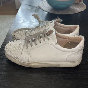 Vita sneakers med nitar - Snygga vita sneakers med nitar på tån och röd sula. Skorna har snörning och en rund tå. Perfekta för att ge din outfit en edgy touch. Väldigt dåligt skick trasig längst fram