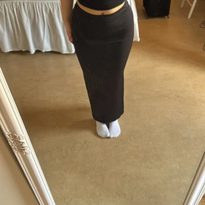 Svart långkjol med slits - Elegant svart långkjol med en stilren slits på sidan. Kjolen har en tight passform som framhäver figuren och är perfekt för en chic look. Den är enkel att matcha med olika toppar för en stilfull outfit.