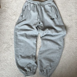 Gråa baggy mjukisbyxor från Poser - Säljer ett par gråa baggy mjukisbyxor från Poser i storlek medium. Byxorna har resår i midjan och vid anklarna, snörning, två fickor fram med dragkedja och en bakficka med logga. Perfekta för en avslappnad stil. Resoren vid anklarna kan även vikas upp för att sitta som ett par vanliga byxor. De är även extra tjocka med 460 gsm.