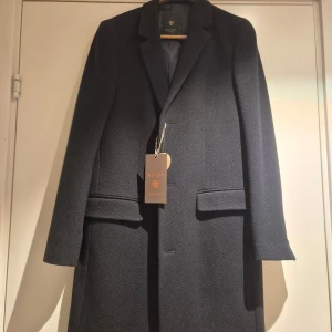 Mörkblå rock från Riley storlek 44 med prislapp kvar - Stilren mörkblå rock från Riley med klassisk krage och knäppning framtill. Jackan har två fickor med lock och två innerfickor med knapp. Helt ny med prislapp kvar. 