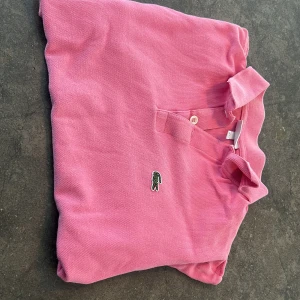 Rosa pikétröja från Lacoste - Säljer en klassisk rosa pikétröja från Lacoste med den ikoniska gröna krokodilloggan på bröstet. Tröjan har korta ärmar, krage och knappar vid halsen. Perfekt för en sportig och avslappnad stil.