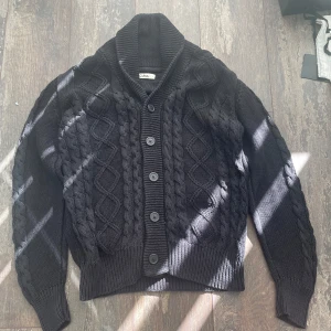 Svart stickad kofta  - Säljer en snygg svart stickad kofta med kabelstickat mönster och knappar framtill. Perfekt för kyliga dagar med sin mysiga och stilrena design. Passar både till jeans och chinos. Storlek: M, Skick: 10/10