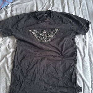 Armani tshirt - Snygg svart t-shirt från Emporio Armani med en glittrig logga på bröstet. Perfekt för att ge din outfit lite extra bling. Kortärmad och bekväm passform.