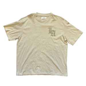 Beige t-shirt med tryck - Snygg beige t-shirt med 'LA' tryck på framsidan och ett konstnärligt motiv på baksidan. Perfekt för en avslappnad stil.