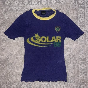 Blå t-shirt , Urban Outfitters - Säljer en blå t-shirt från Urban Outfitters med gult tryck 'SOLAR 96' och ett emblem på bröstet. T-shirten har en gul krage och är kortärmad. 💜 använd bara en gång 