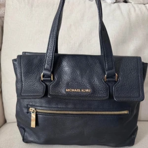 Svart handväska från Michael Kors - Elegant svart handväska från Michael Kors i skinn med guldfärgade detaljer. Väskan har en praktisk dragkedja framtill och dubbla handtag för enkel bärning. Perfekt för att hålla dina saker organiserade och stiliga.
