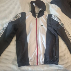 Lacoste sport jacka - Vit och svart vindjacka från Lacoste sport med huva och dragkedja i rött. Jackan har långa ärmar med Lacoste-logga längs sidorna. 