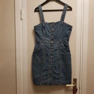 Jeans hängsel klänning med knappar strl 38 - Snygg jeansklänning med knappar framtill och praktiska fickor. Klänningen har breda axelband (hängslen) och en figurnära passform. Perfekt för en avslappnad stil med en touch av denim. Det står 40 men är en 38.
