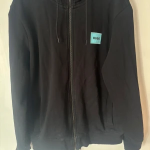 Svart hoodie med dragkedja från Hugo - Säljer en svart hoodie från Hugo med dragkedja framtill och en ljusblå patch med logga på bröstet. Tröjan har huva med snörning och ribbade muddar vid ärmslut och nederkant. Perfekt för en avslappnad stil.