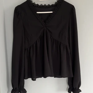 Svart blus med spetsdetaljer - Säljer en svart blus med v-ringning och fina spetsdetaljer runt halsen. Blusen har långa ärmar med resår vid ärmslut och ett löst, flowy fall. Perfekt för dig som gillar stilrena och feminina plagg.