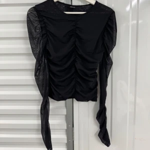Svart mesh topp med rynkade detaljer - Säljer en helt ny svart mesh topp med långa, transparenta ärmar och rynkade detaljer framtill. Toppen har en tight passform och är perfekt för att skapa en trendig look.