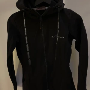 Svart hoodie från Peak Performance med dragkedja och huva - Snygg svart hoodie med dragkedja och huva. Jackan har långa ärmar och snören med textdetaljer. 