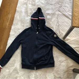  Moncler Hoodie  - Säljer en mörkblå hoodie från Moncler med dragkedja och huva. Tricolore-detalj på huvan och logotyp på ärmen. Perfekt för en sportig och stilren look. Fickor framtill och ribbade muddar. Helt ny skick nästan