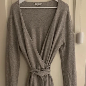 Grå omlottklänning i kashmir från Davida -  Grå omlottklänning från Davida i 100% kashmir. Klänningen har lång ärm, knytskärp i midjan och djup v-ringning. Finns två små mål på framsidan vid låret men syns inte och går att sy. Klänningen går till en bit över knäet