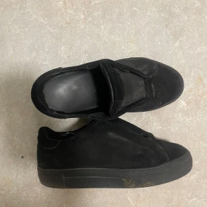 Svarta mocka sneakers - Säljer ett par helsvarta sneakers i mocka med platt sula och rund tå. Skorna har en minimalistisk design utan synlig snörning och är perfekta för en clean look. Passar dig som gillar stilrena och enkla skor.