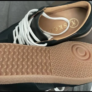 Svarta sneakers från Skolyx med vit sula - Snygga svarta sneakers från Skolyx med vit snörning och beige yttersula. Skorna har en klassisk design med rund tå och paneler i olika material för en stilren look. Perfekta för dig som gillar en clean och tidlös stil.